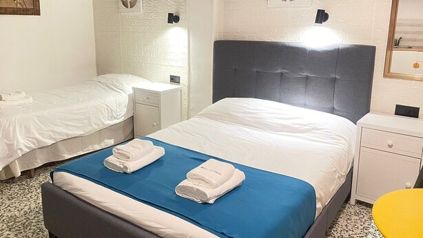 Et hotelværelse med to senge, en stor grå sengegavl og et lille natbord.