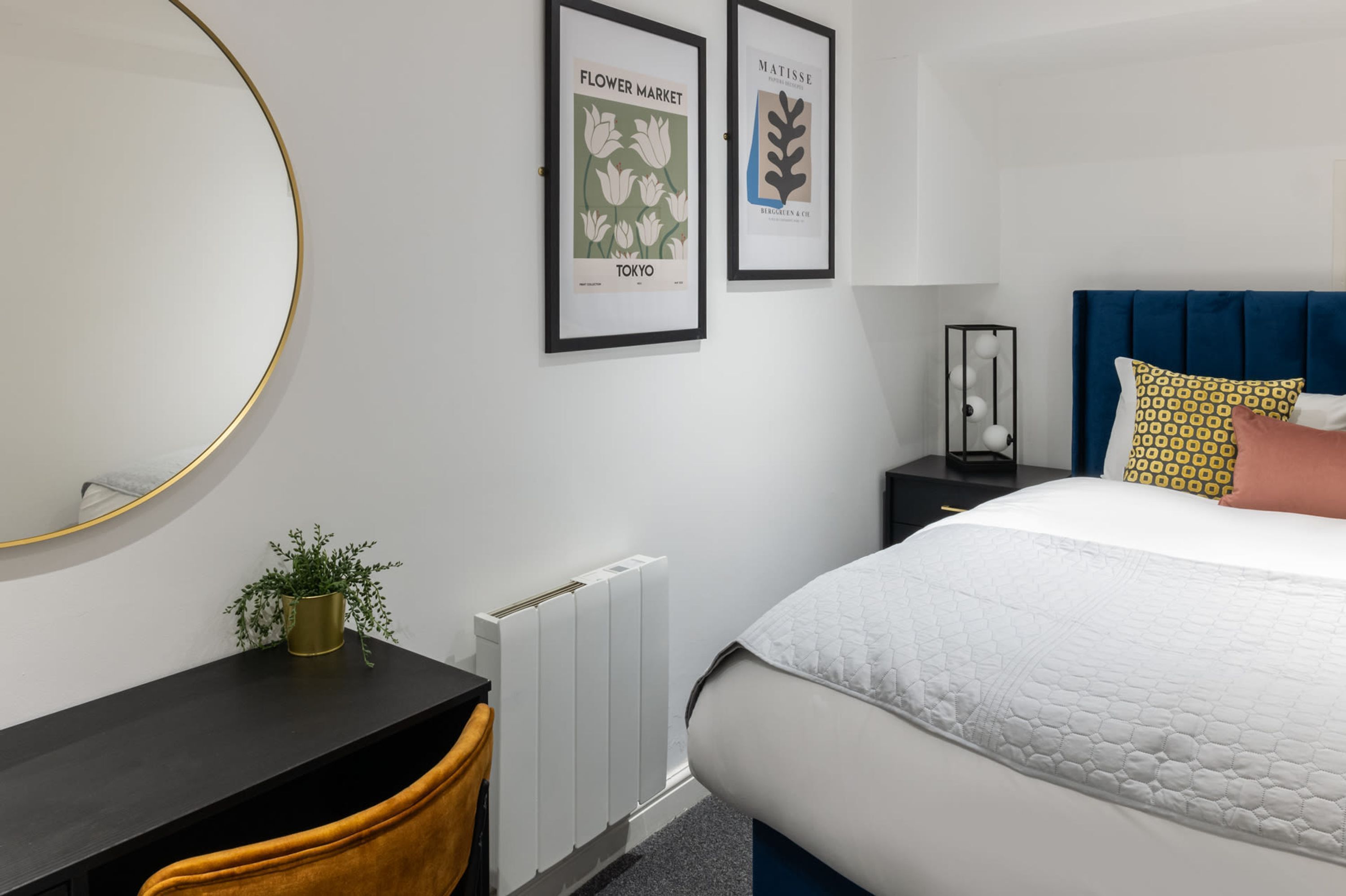 Ein Schlafzimmer mit einem Bett, einem Nachttisch mit Pflanze, einer Heizung und zwei gerahmten Bildern an der Wand.