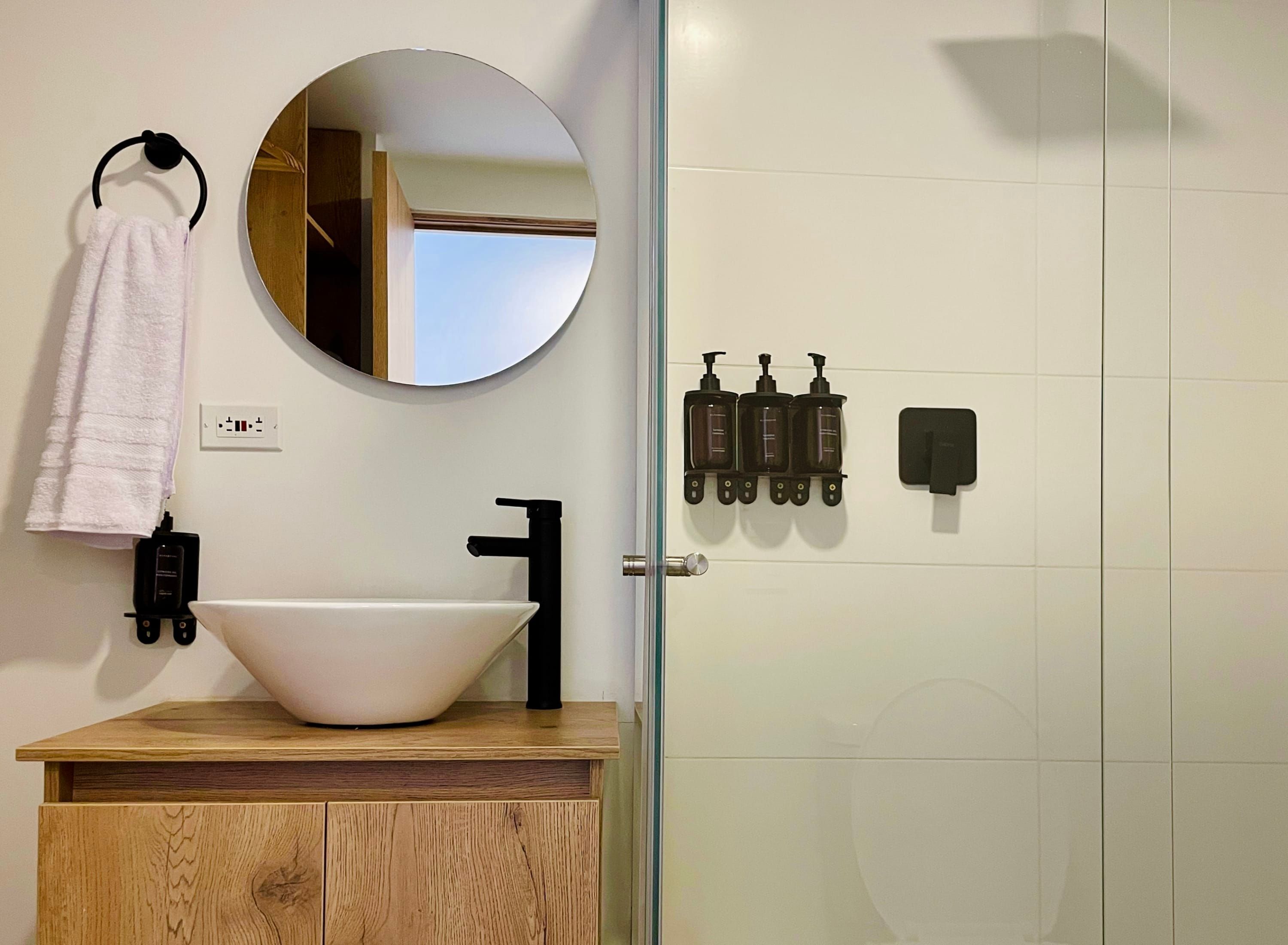 Une salle de bain avec un lavabo blanc, un robinet noir et un miroir rond au-dessus. Une serviette est accrochée à un anneau noir, il y a une prise de courant et un ensemble de distributeurs de savon noirs fixés au mur.