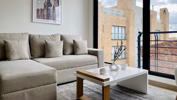 Un salon moderne avec un canapé beige, une table basse blanche et une grande fenêtre donnant sur des immeubles en briques.