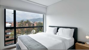 Une chambre d’hôtel avec une grande fenêtre, un lit recouvert de draps blancs, une tête de lit sombre et une table de chevet avec une lampe.
