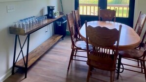 A dining area with a round table and wooden chairs, a kitchen counter with a coffee maker, and a view of a lake through large windows. - Camp Blue Canoe - Creekside Cottage  (Three Rivers)