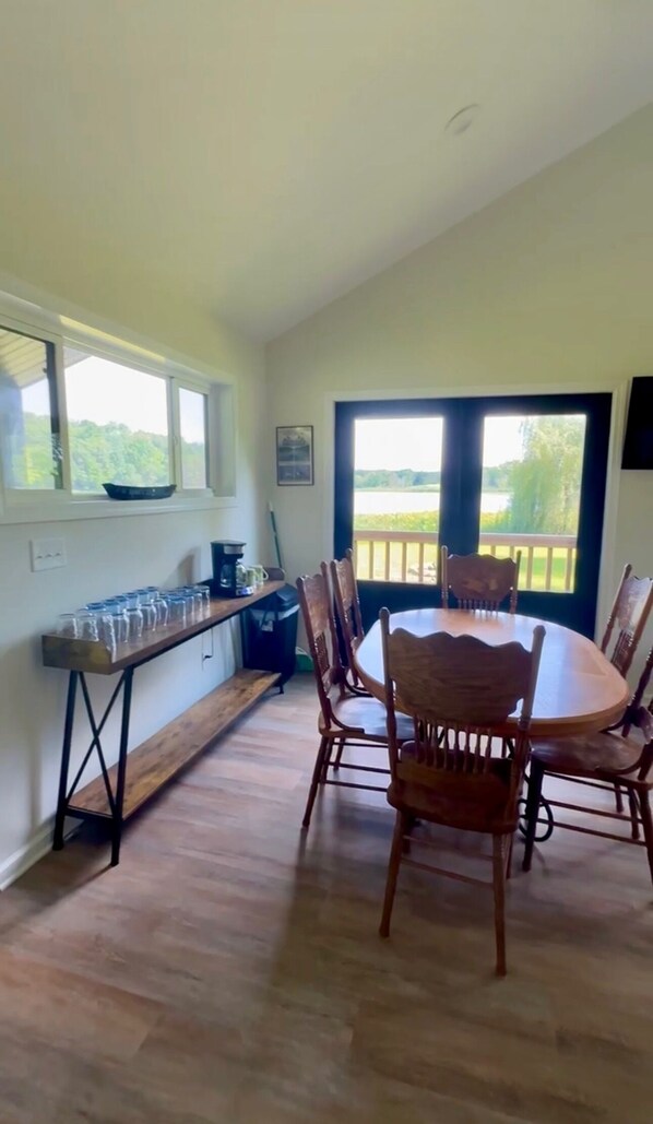A dining area with a round table and wooden chairs, a kitchen counter with a coffee maker, and a view of a lake through large windows.