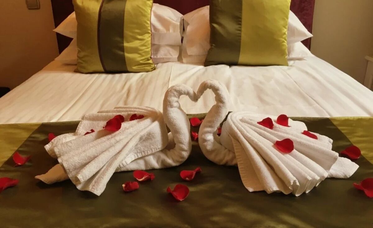 A bed with a white bedspread, two towels shaped like swans, and red rose petals scattered on the bed.