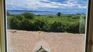 A view through a window with a gravel path, greenery, and a distant body of water under a partly cloudy sky.