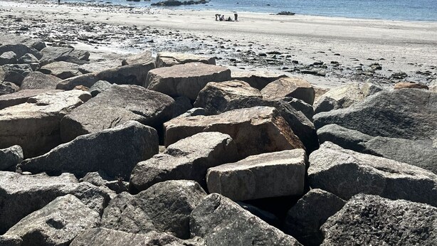 A rocky beach with large boulders in the foreground and a clear view of the sea and distant hills.