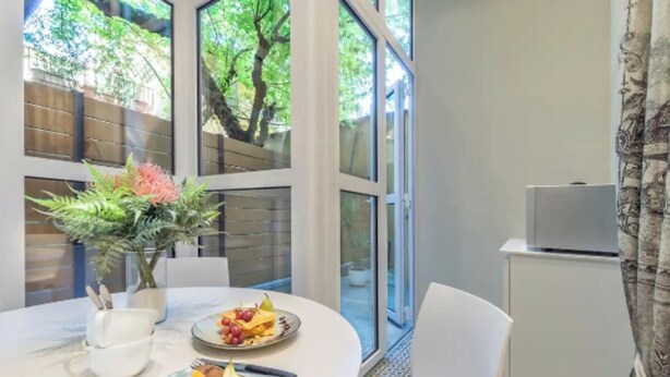 Un comedor con una mesa redonda, sillas blancas, una planta en maceta y vistas al exterior con vegetación.