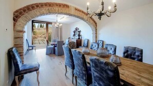 A dining area with a wooden table, upholstered chairs, and a chandelier. An arched brick structure frames the entrance to another room. - Villa Blu - Seven Bedroom Villa, Sleeps 14 (Greve in Chianti)