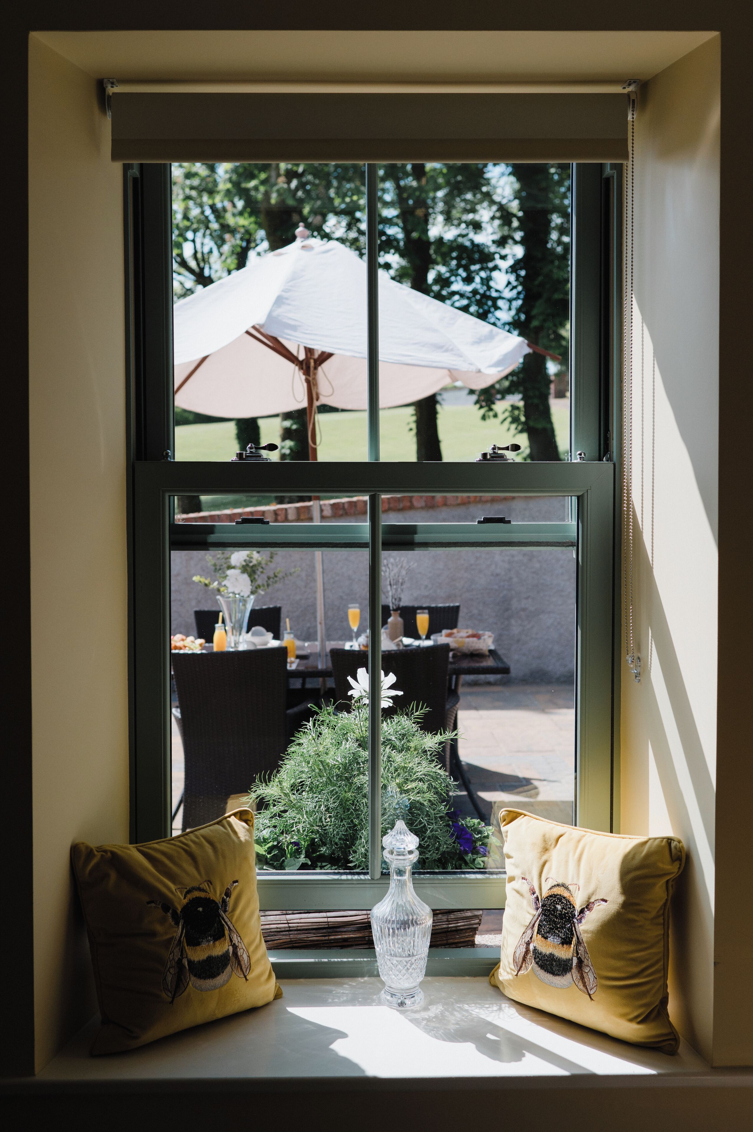 A window with a view of an outdoor dining area, a table with a vase and a bottle, and two pillows with bee designs.