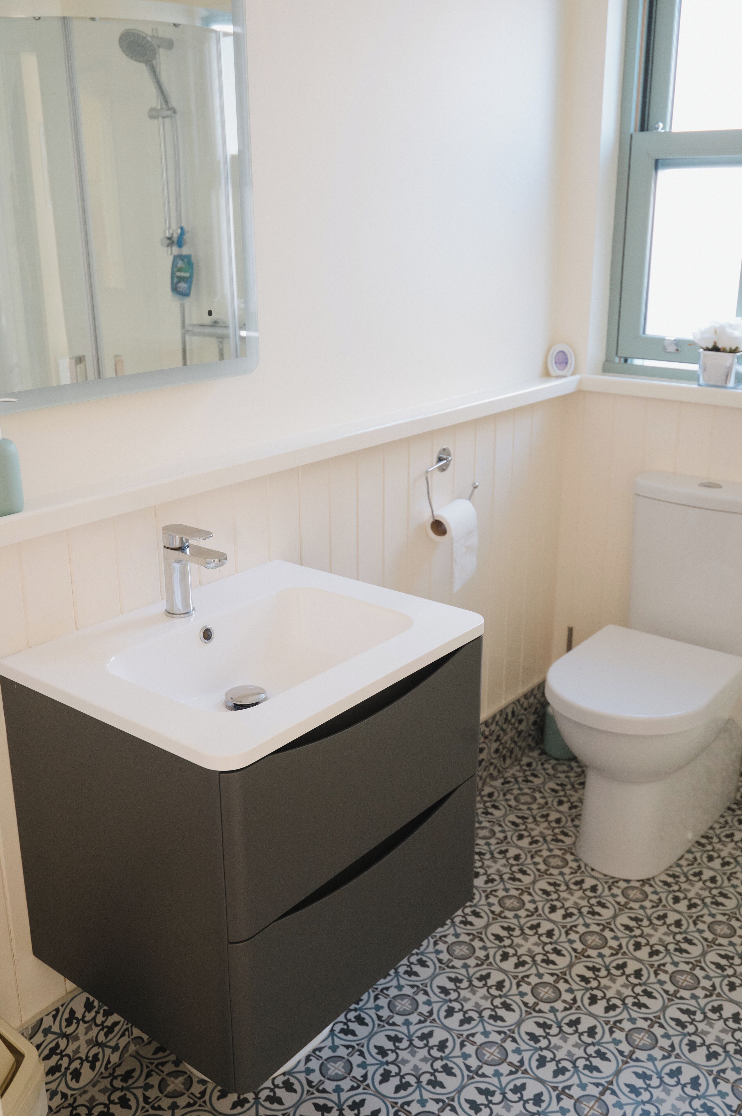 A bathroom with a white sink, dark cabinet, toilet, patterned floor, and a window with a view.