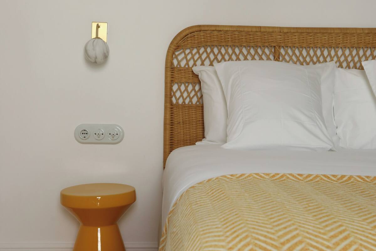 A bedroom with a wicker headboard, white bedding, a patterned throw, a small orange stool, and a wall-mounted light fixture.