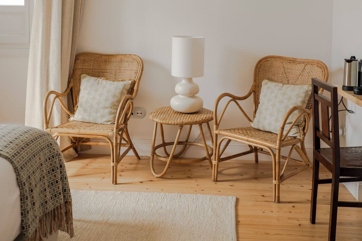 A cozy room with two wicker chairs, a small table with a lamp, a bed with a blanket, and a painting on the wall.