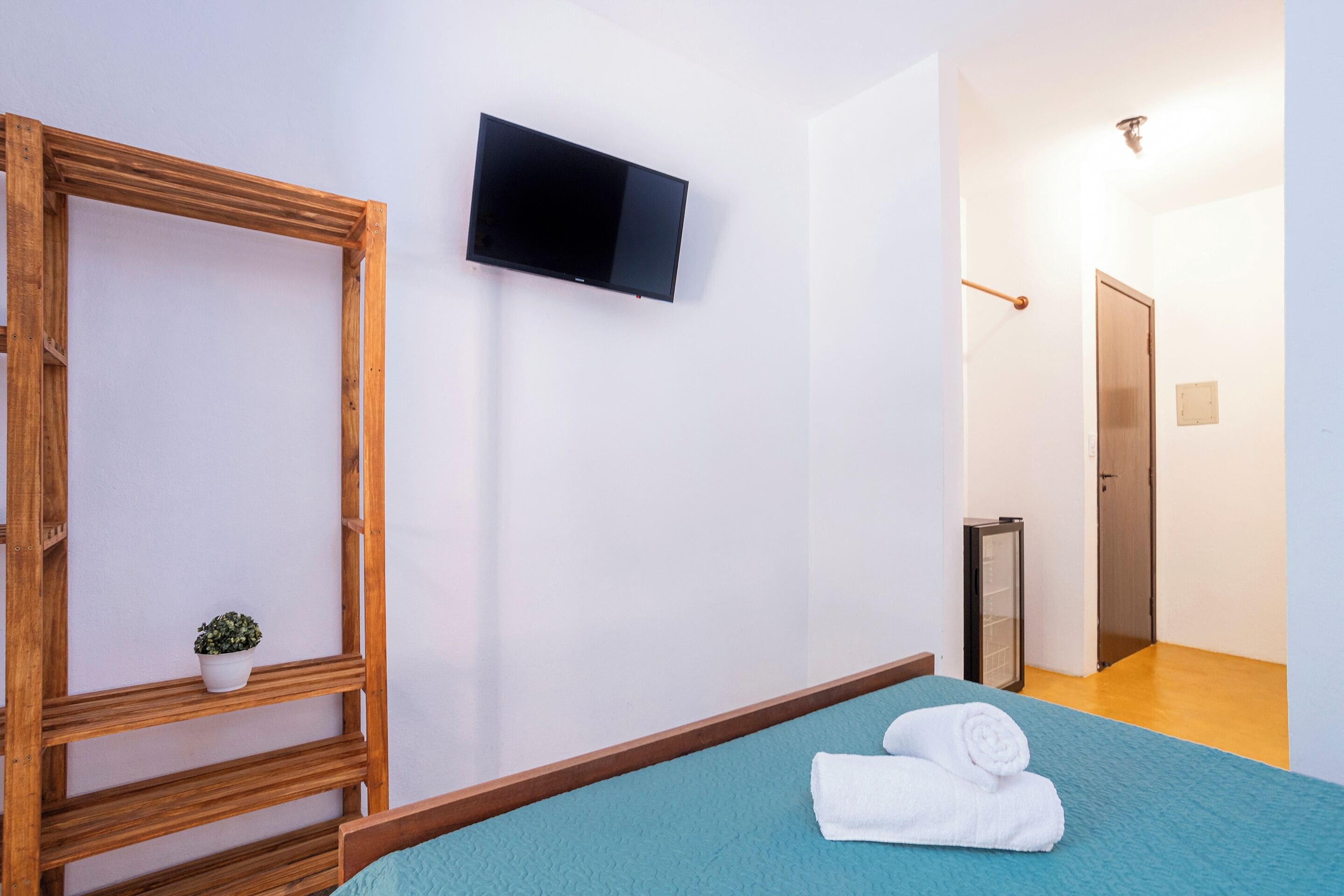 Een kamer met een bed, een aan de muur bevestigde televisie, een houten frame, een kleine potplant en een deur naar een andere kamer.
