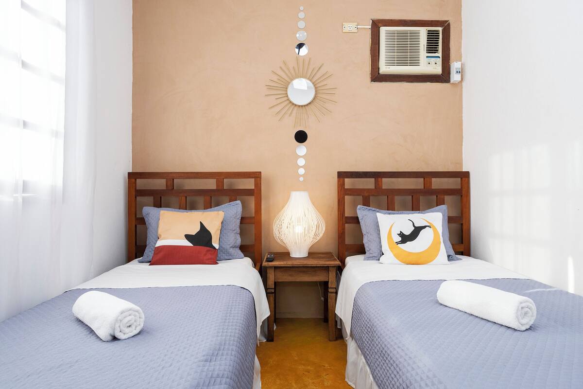 A hotel room with two beds, a nightstand with a lamp, a wall-mounted air conditioner, and a decorative sun mirror.