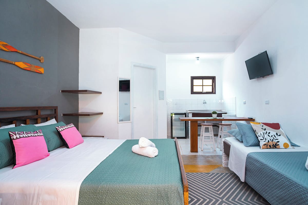 A modern hotel room with a bed, a sofa, a small kitchen area, and a TV mounted on the wall.