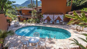 A pool area with lounge chairs, a table, and a pergola with white drapes. - Pousada Aysu Maresias (São Sebastião)