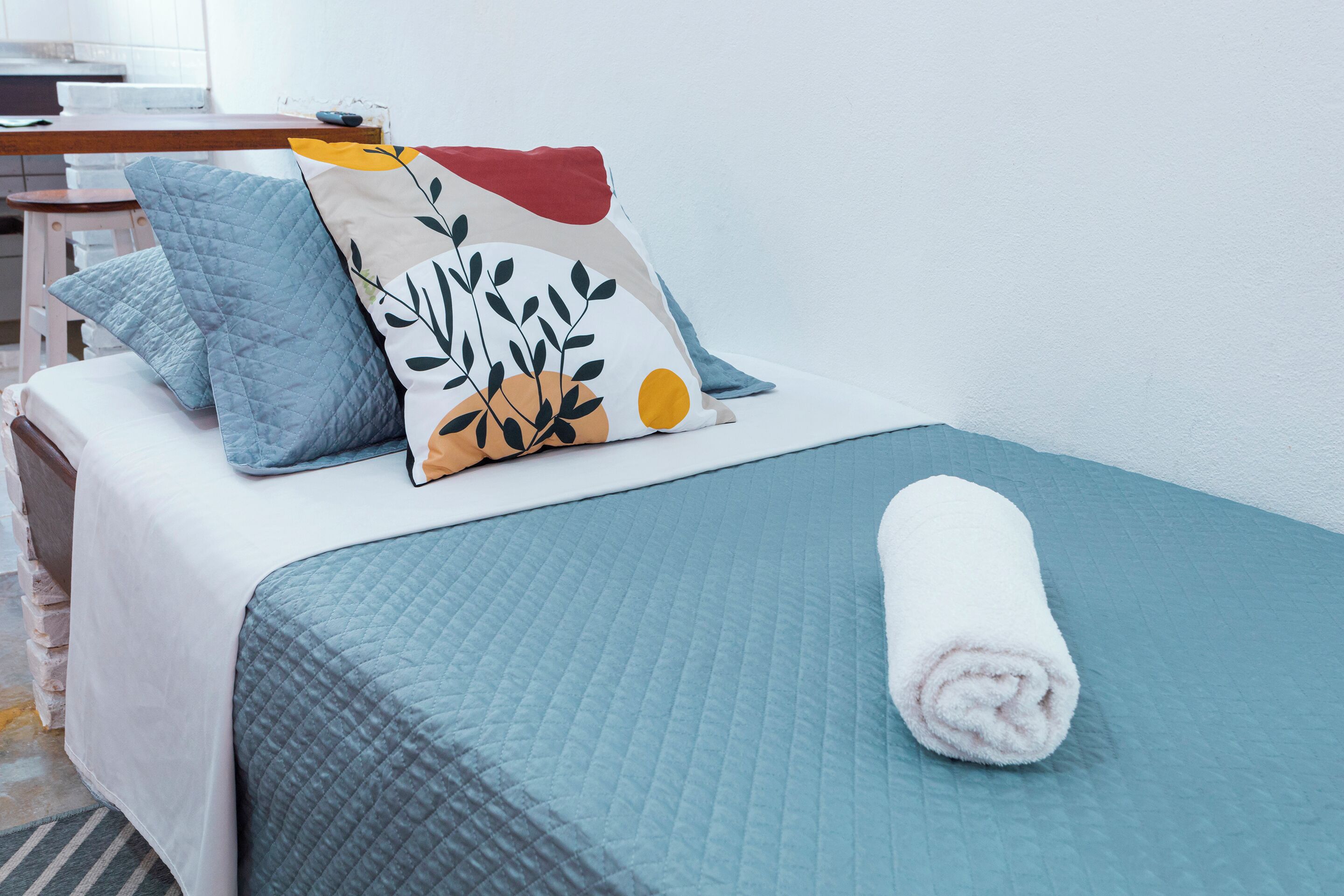 Una cama bien hecha con un edredón azul, una toalla blanca enrollada y dos almohadas con diseño de hojas.