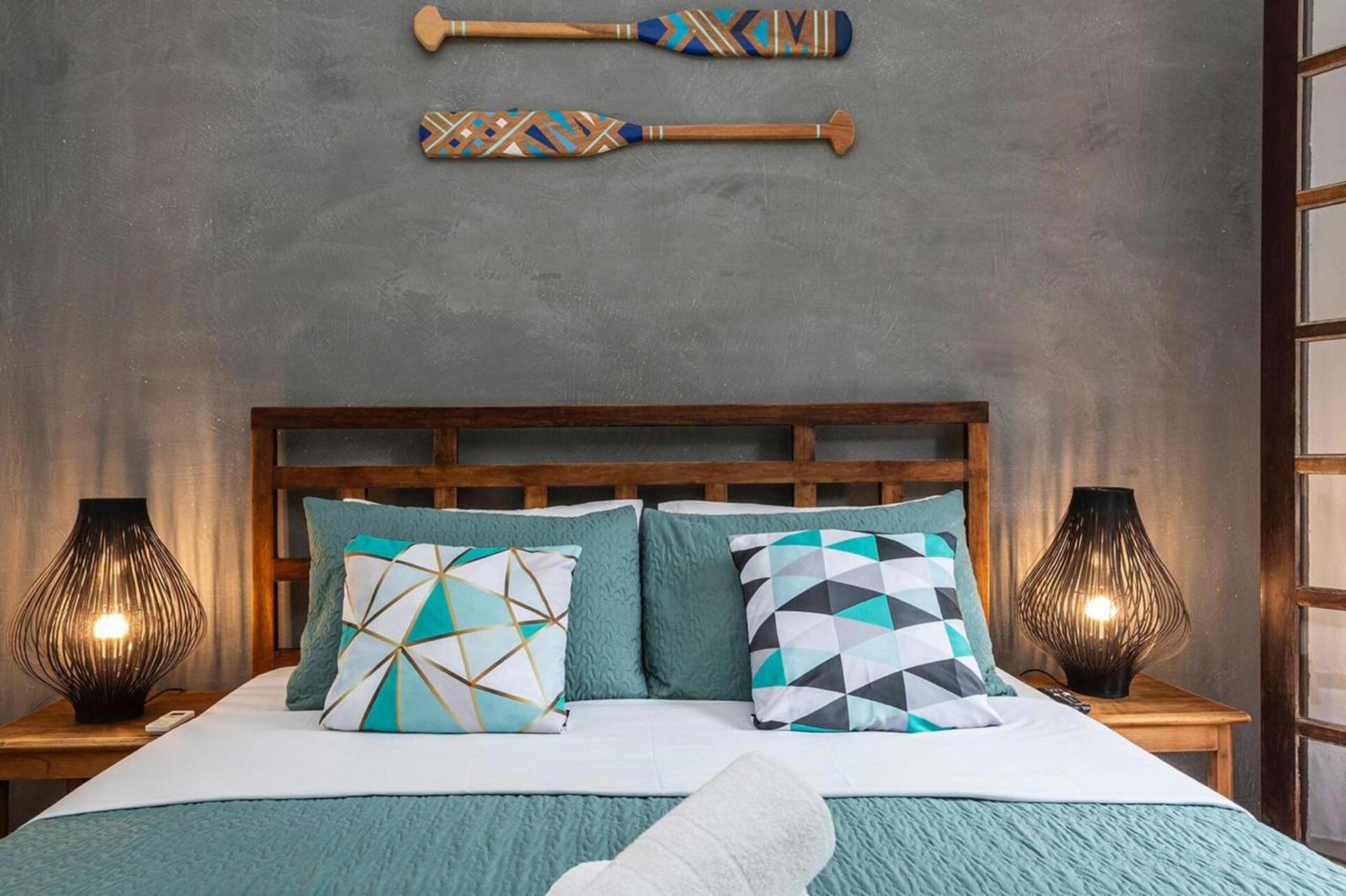 Un lit bien fait, agrémenté d’oreillers à motifs, d’une tête de lit en bois et de deux lampes fixées au mur, une de chaque côté.