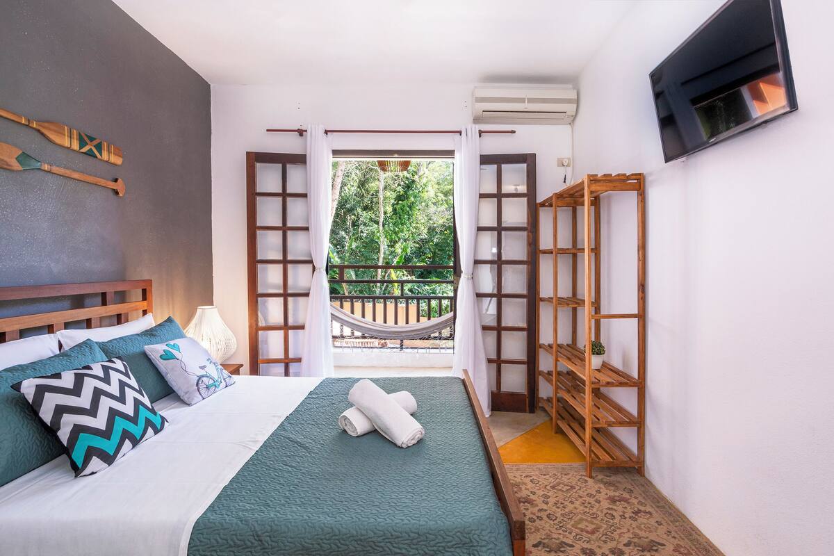 A bedroom with a bed, pillows, a wooden shelf, and a view of a balcony with greenery.