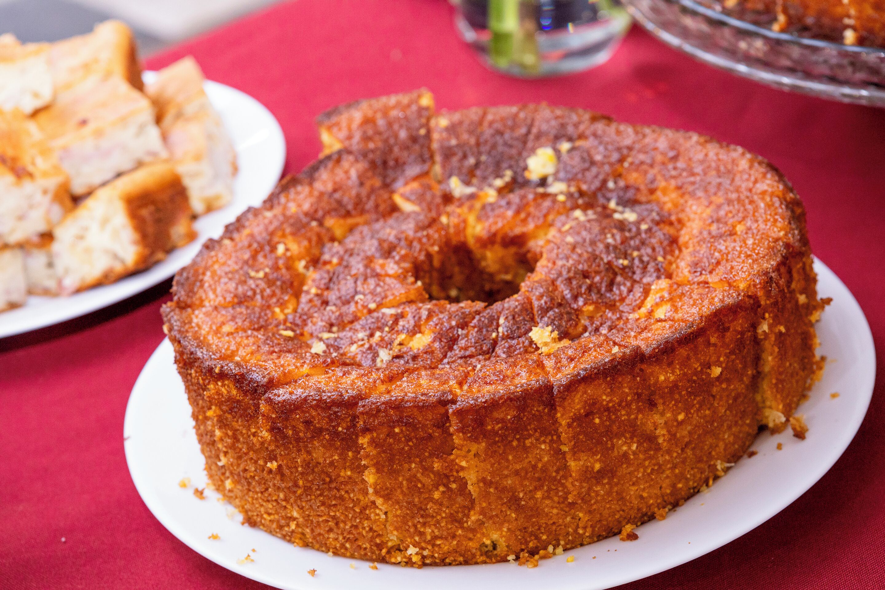A round cake on a white plate with crumbs around it, placed on a red tablecloth.