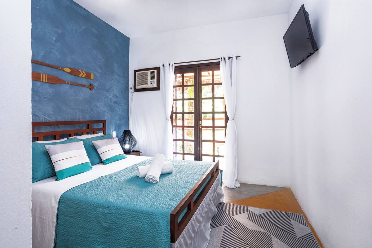 A bedroom with a bed, a window with curtains, a wall-mounted TV, and a small air conditioning unit.