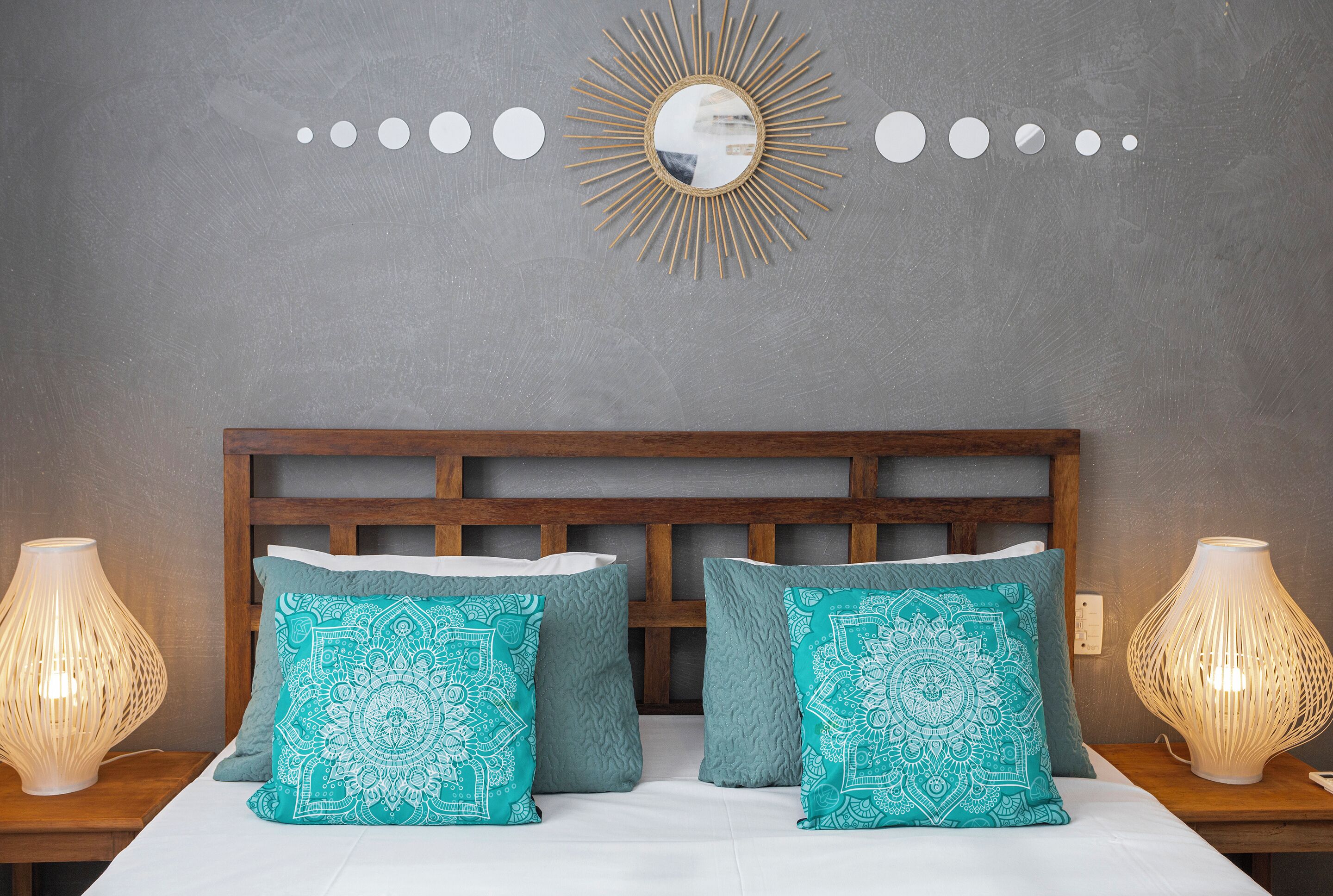 Una cama bien hecha con dos almohadas estampadas, un cabecero de madera y un espejo montado en la pared con diseño de sol.