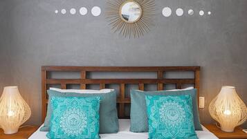 Un lit bien fait, agrémenté de deux coussins à motifs, d’une tête de lit en bois et d’un miroir mural orné d’un motif en forme de soleil.