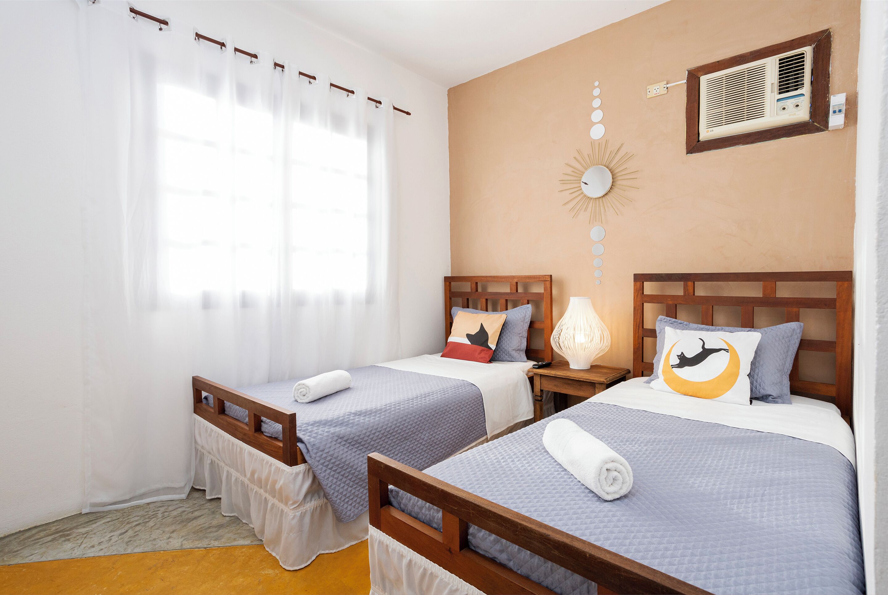 A hotel room with two single beds, a wooden headboard, a wall-mounted air conditioner, a sunburst decoration, and a window with sheer curtains.
