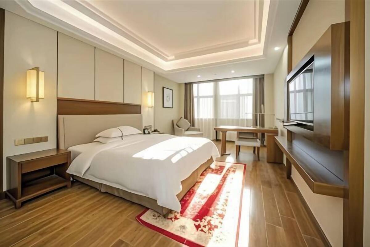 Habitación de hotel con cama, mesita de noche, televisor, escritorio, silla y un ventanal grande.