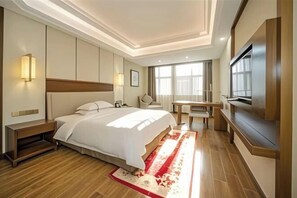 Habitación de hotel con cama, mesita de noche, televisor, escritorio, silla y un ventanal grande.