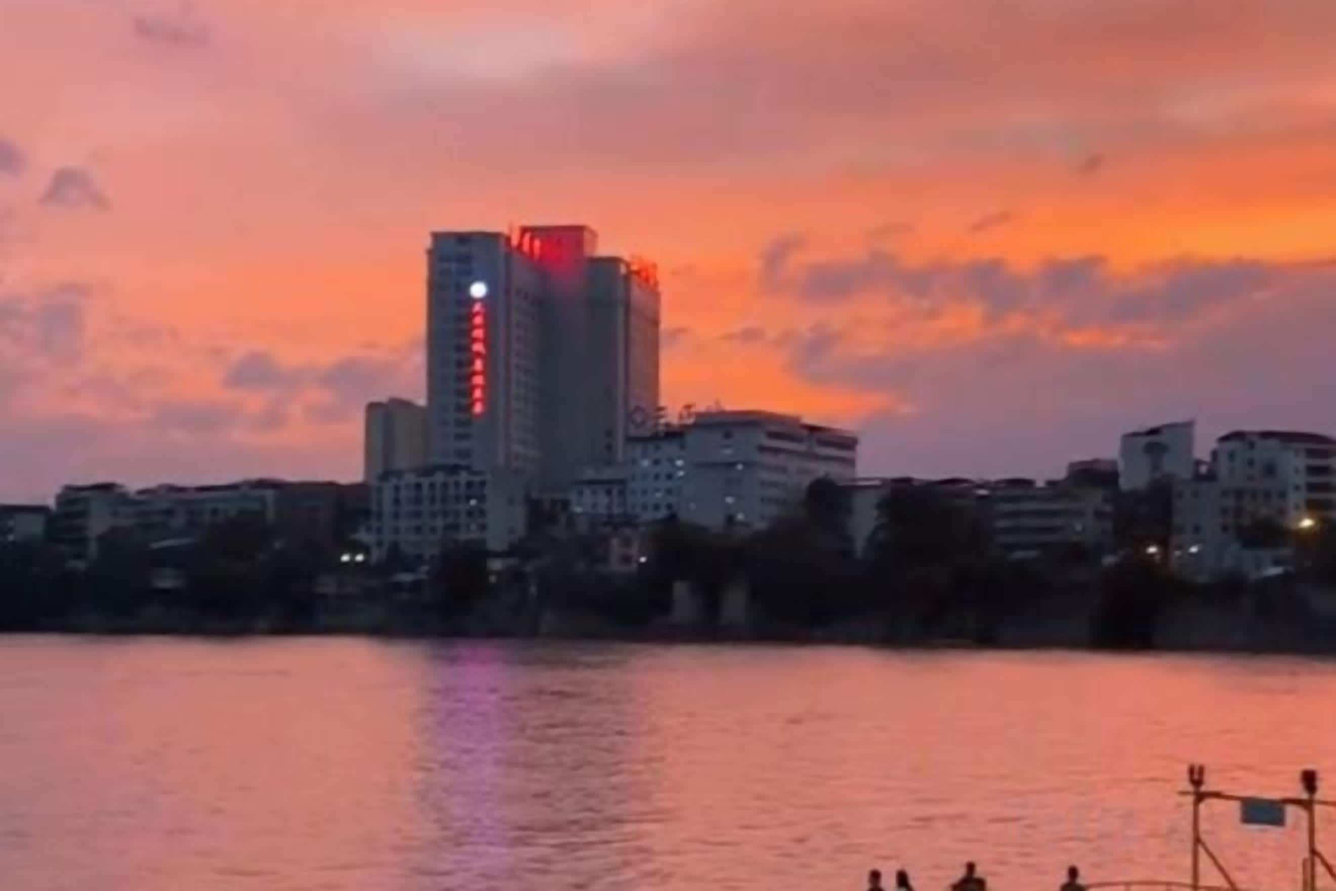A cityscape with a prominent building with red lights at the top, a body of water in the foreground, and other buildings in the background.