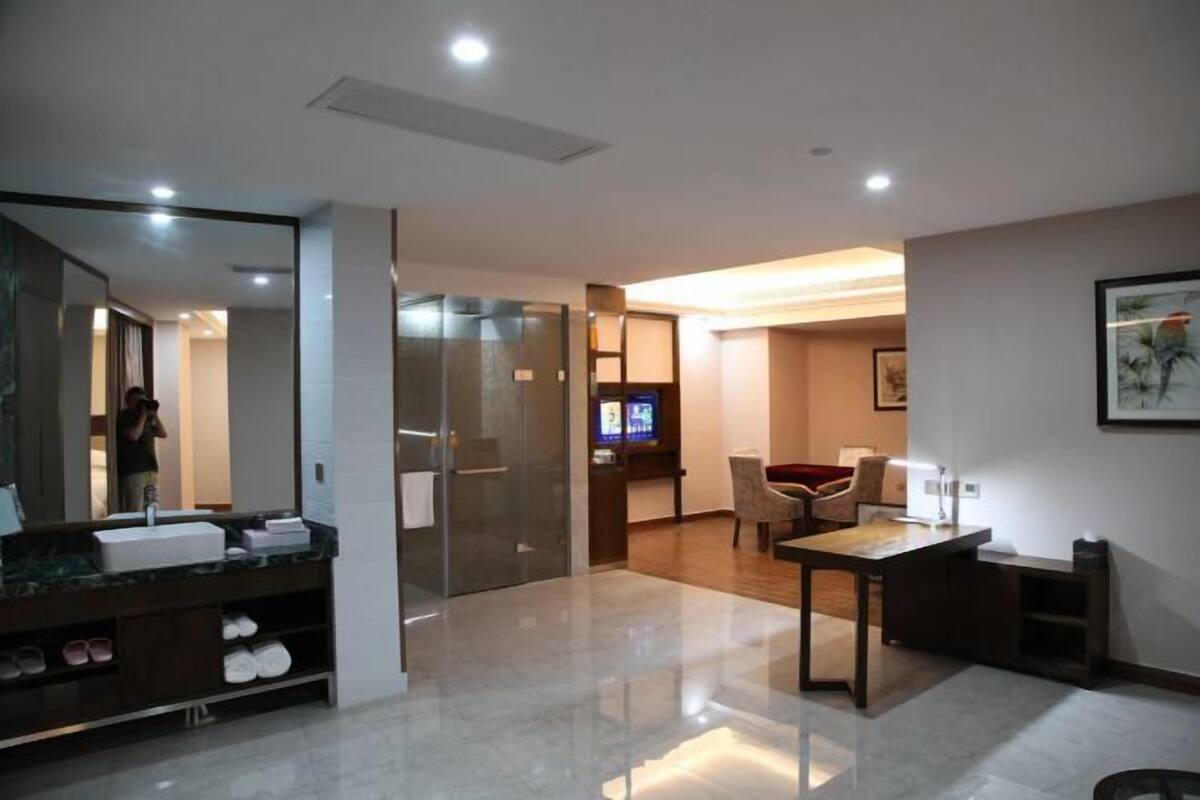 Una habitación de hotel moderna con un espejo grande, un lavamanos, una mesa de madera y un televisor al fondo.