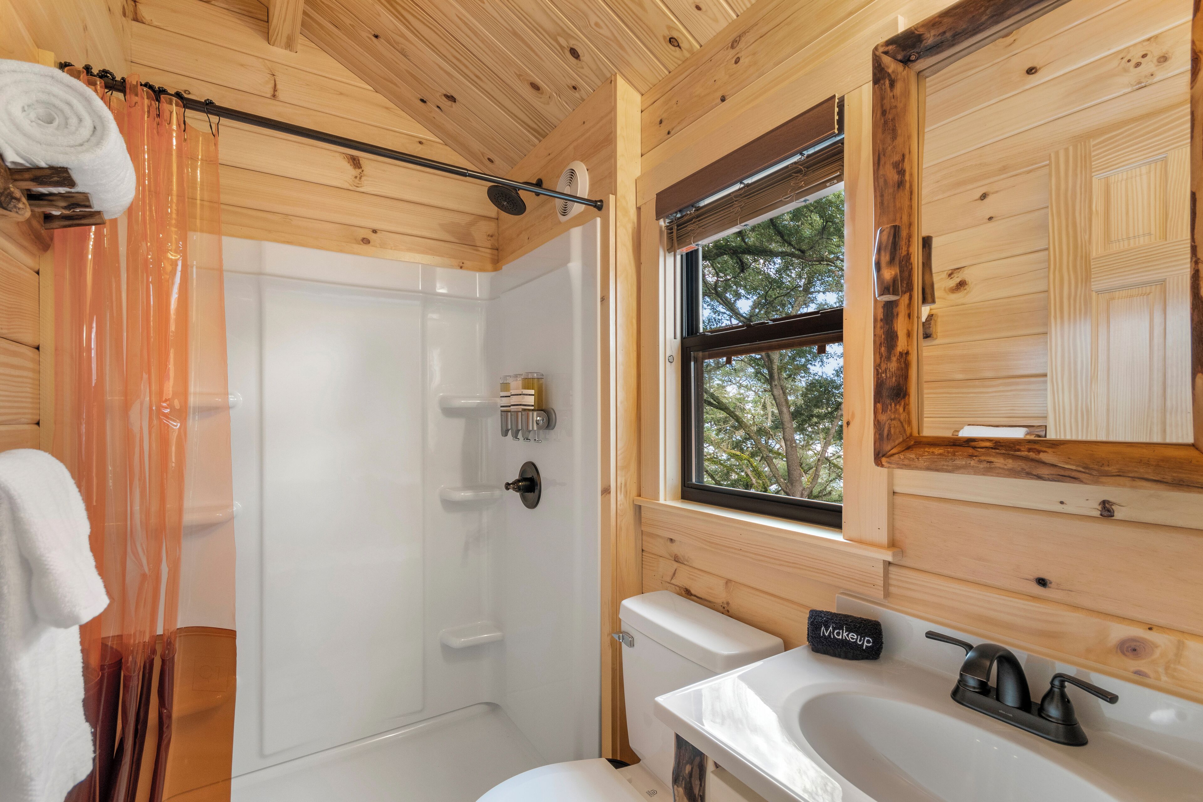 A wooden cabin bathroom with a shower, toilet, sink, and a window with a view of trees.
