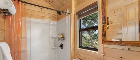 A wooden cabin bathroom with a shower, toilet, sink, and a window with a view of trees.