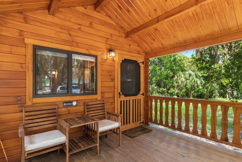 Una cabaña de madera con un porche, dos sillas de madera, una mesa y un ventanal con vistas a un bosque.