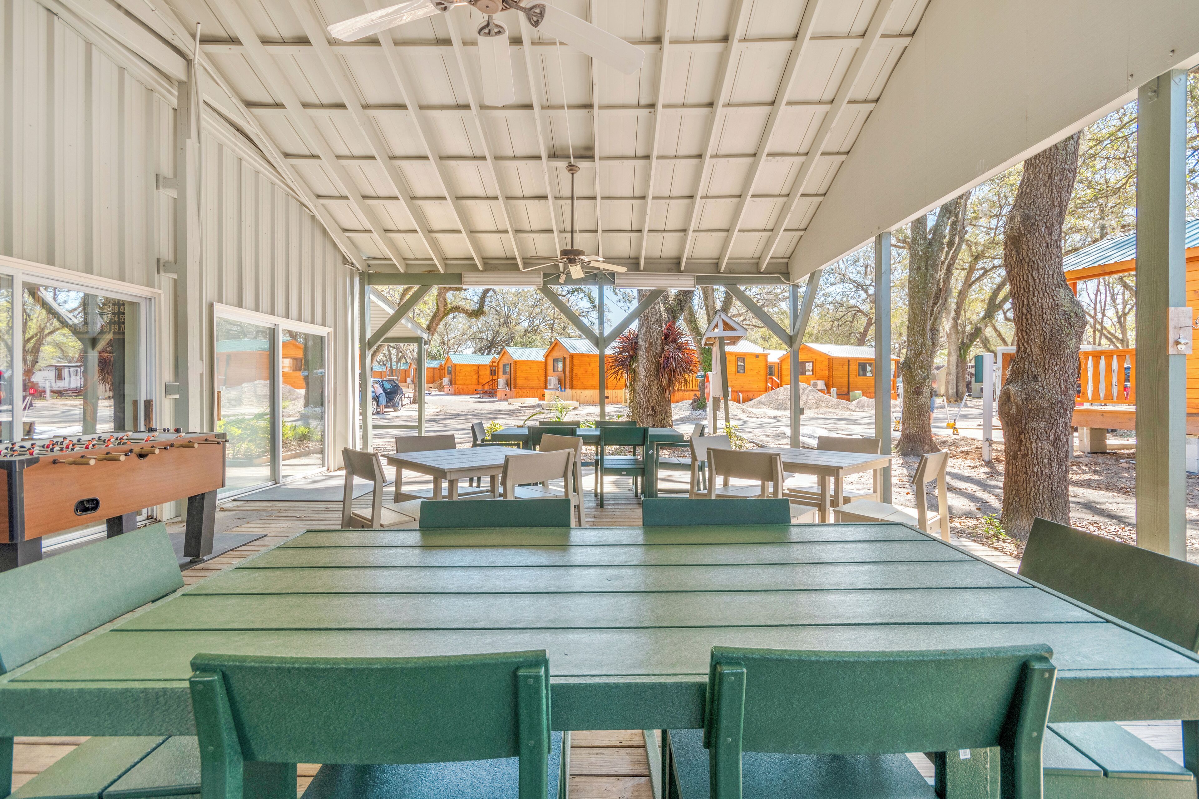 Ein Außenbereich mit einer Terrasse, grünen Tischen und Stühlen, einem Kicker und Blick auf nahegelegene Blockhütten.