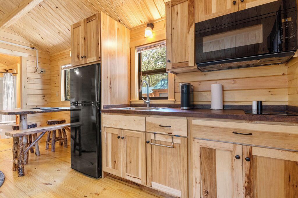 A wooden cabin interior with a kitchen area featuring a black refrigerator, wooden cabinets, a countertop, and a window providing natural light.
