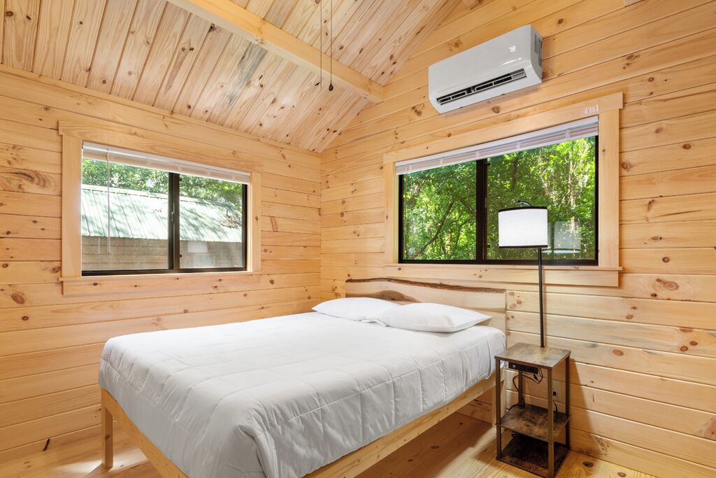 Une pièce de type cabane en bois, comprenant un lit, deux fenêtres et un appareil de climatisation.