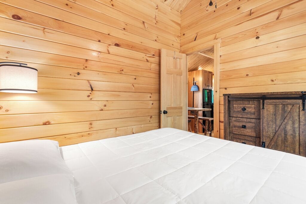 A wooden cabin room with a bed, a wall-mounted lamp, and a rustic fireplace.