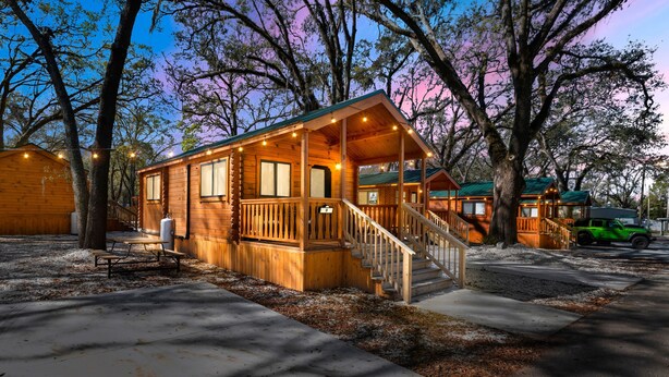A wooden cabin with a porch, a staircase, and a green vehicle parked nearby. - Happy Travelers RV Resort (Thonotosassa)