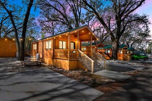 A wooden cabin with a porch, a staircase, and a green vehicle parked nearby. - Happy Travelers RV Resort (Thonotosassa)
