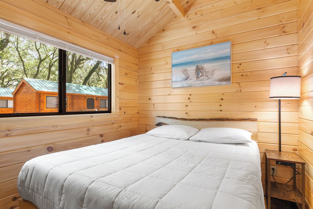 A wooden cabin room with a bed, a window, and a painting of a beach scene.