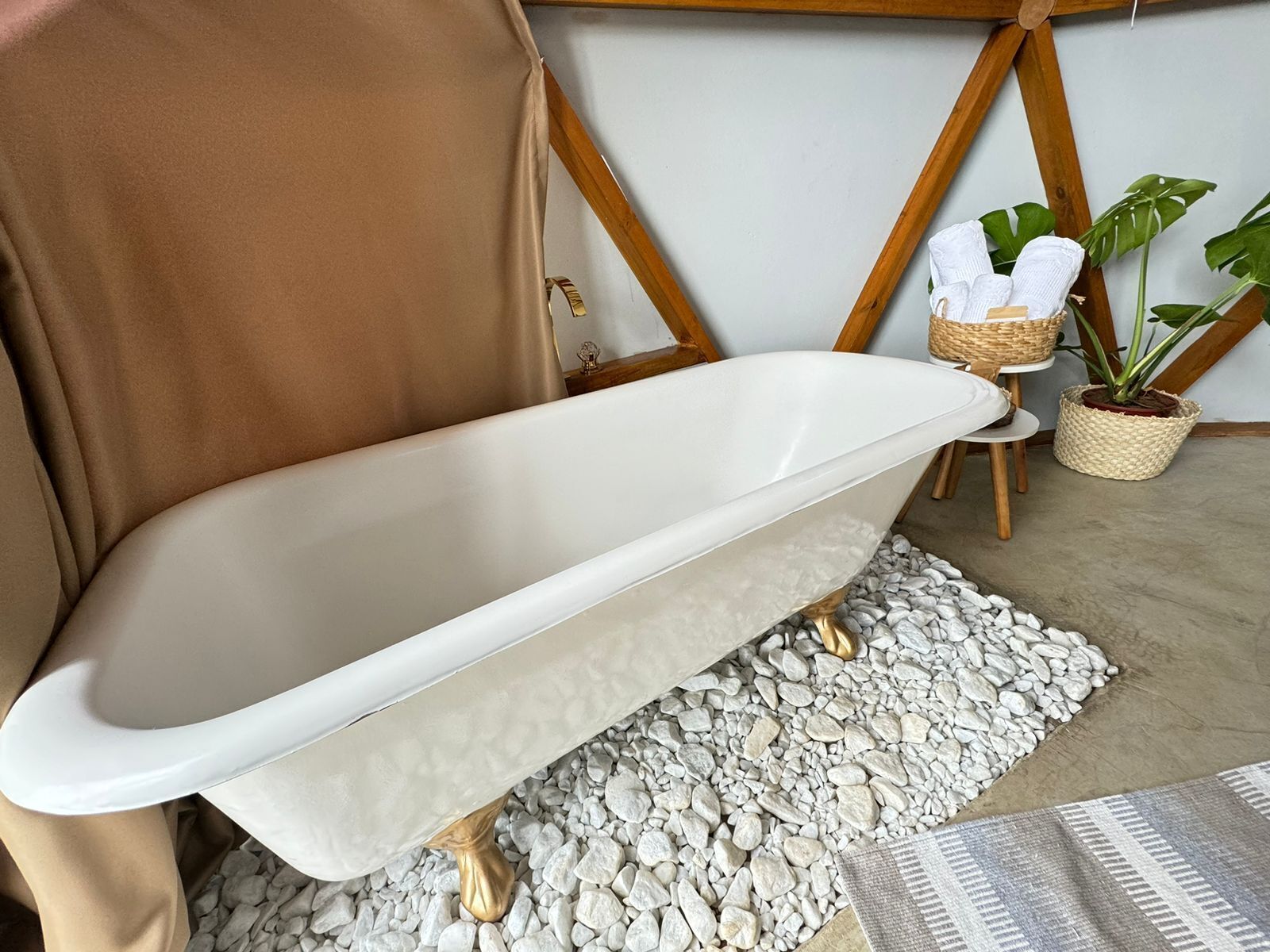 Una bañera con patas doradas, una toalla color beige y un tapete de baño blanco sobre una superficie de guijarros.