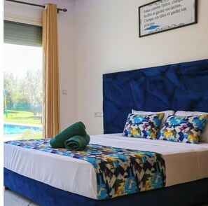 Relax in a spacious room with a large bed, featuring a comfortable headboard and vibrant bedspread, and enjoy a view of the surrounding green landscape.