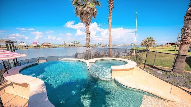 Une piscine au design incurvé, entourée d’une terrasse et de palmiers, offrant une vue sur une étendue d’eau et des bâtiments éloignés.