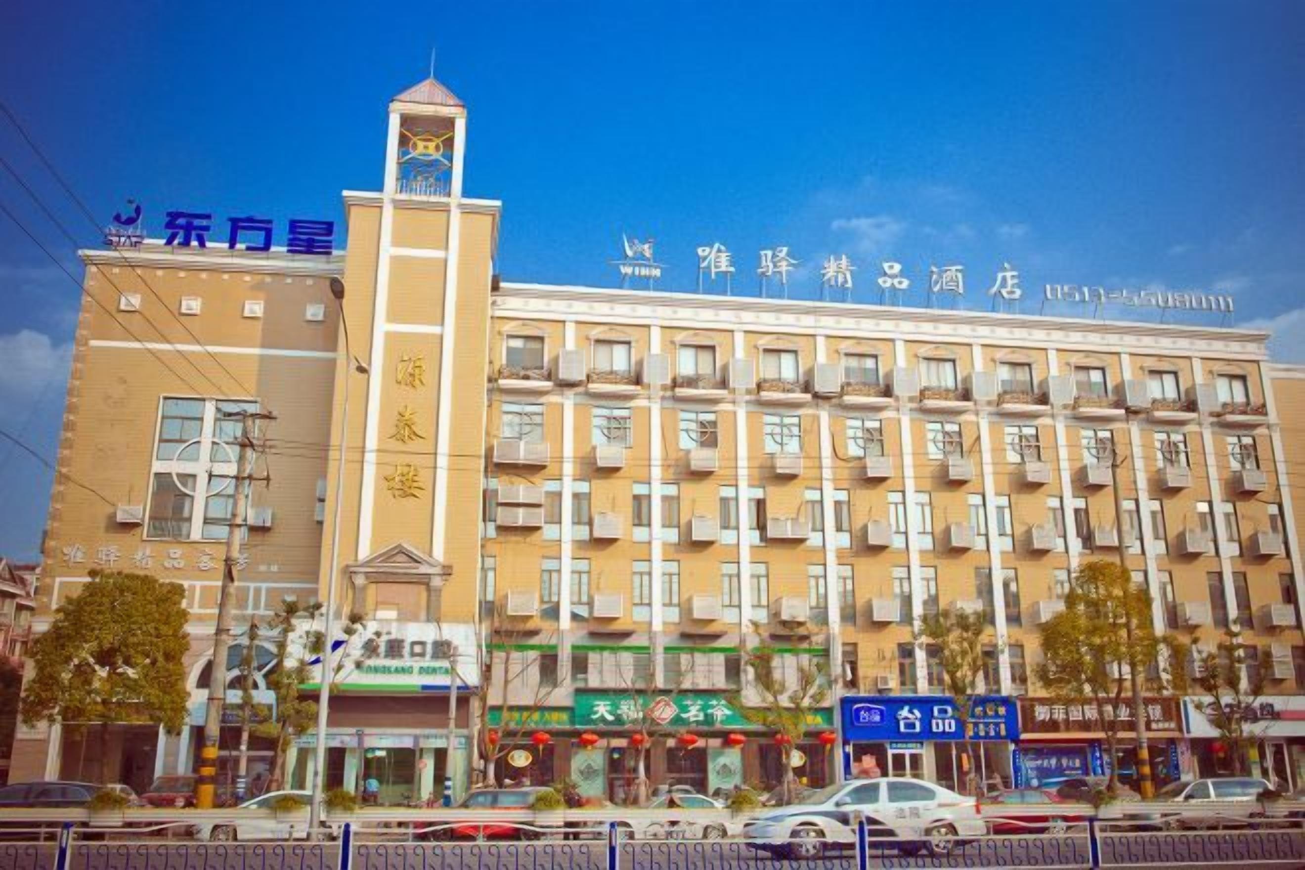 Un hotel de varios pisos con una imponente torre de reloj, varios balcones y una variedad de locales comerciales en la planta baja.