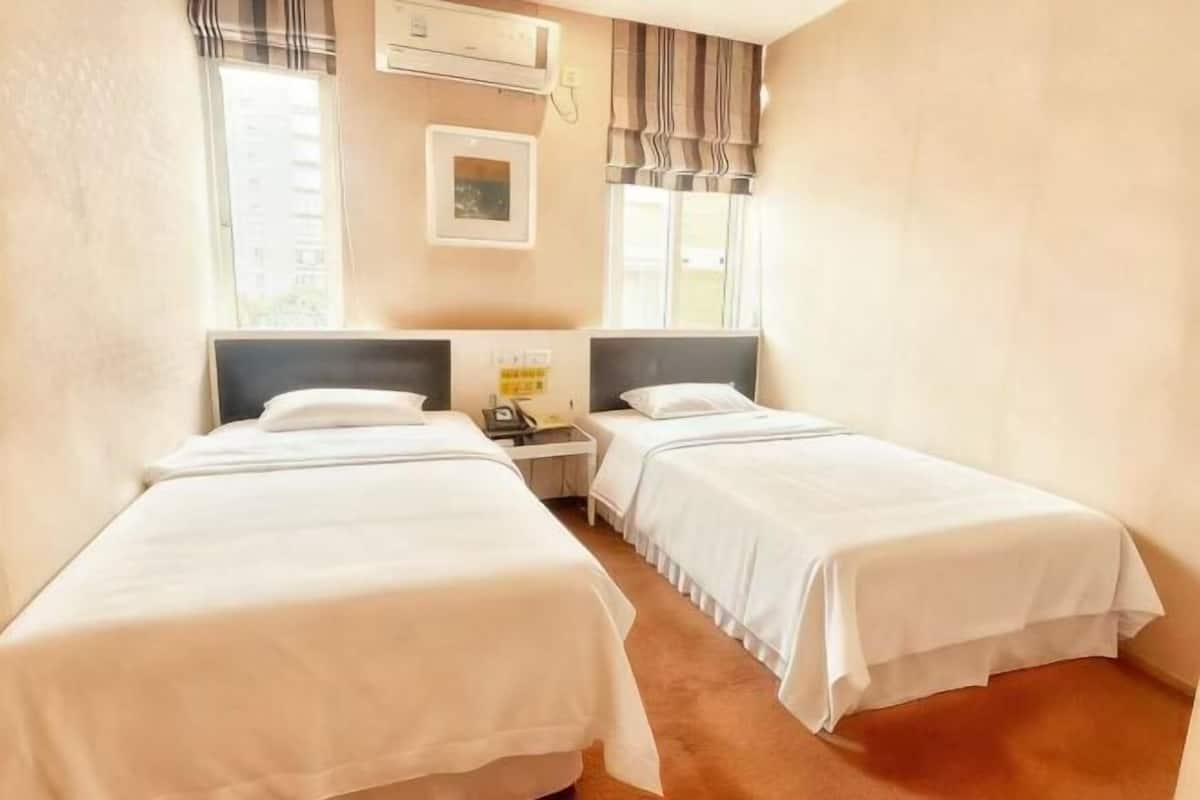 Une chambre d’hôtel avec deux lits simples, une table de chevet avec un téléphone, un climatiseur fixé au mur et une fenêtre avec des rideaux.