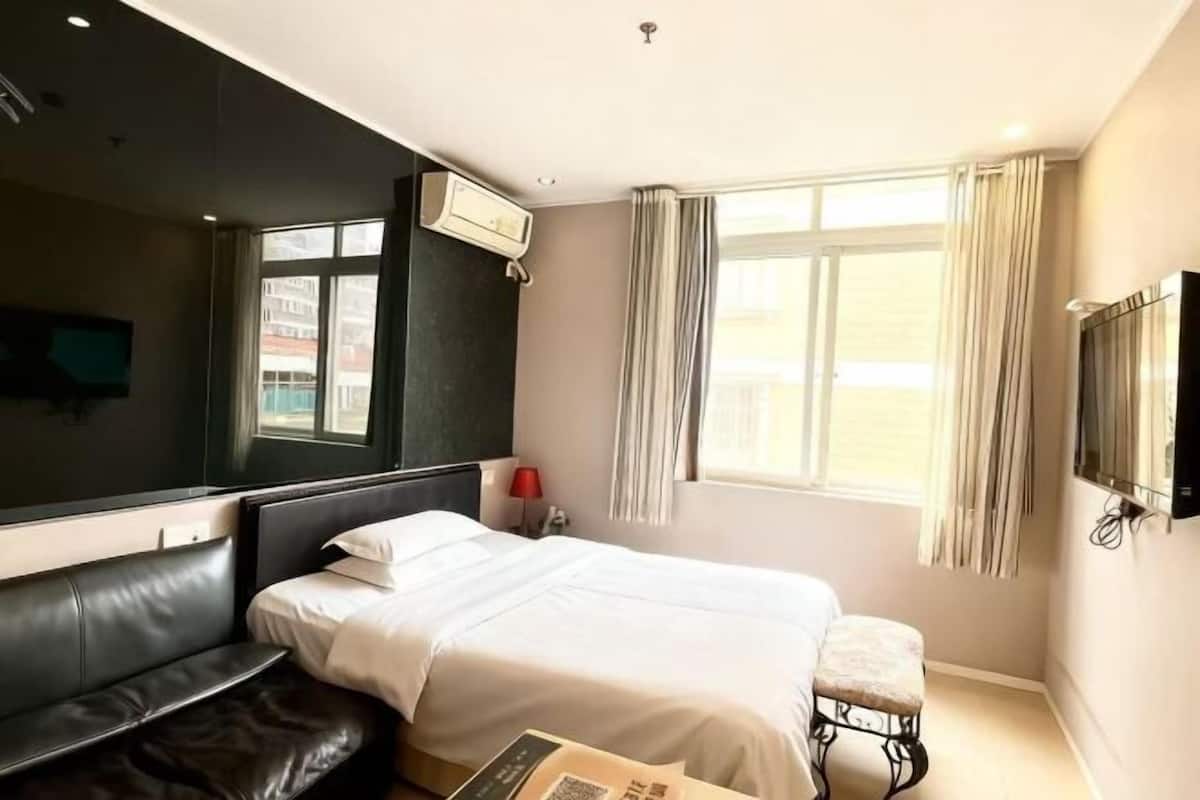Une chambre d’hôtel avec un lit, un petit tabouret, une télévision et une fenêtre avec des rideaux.