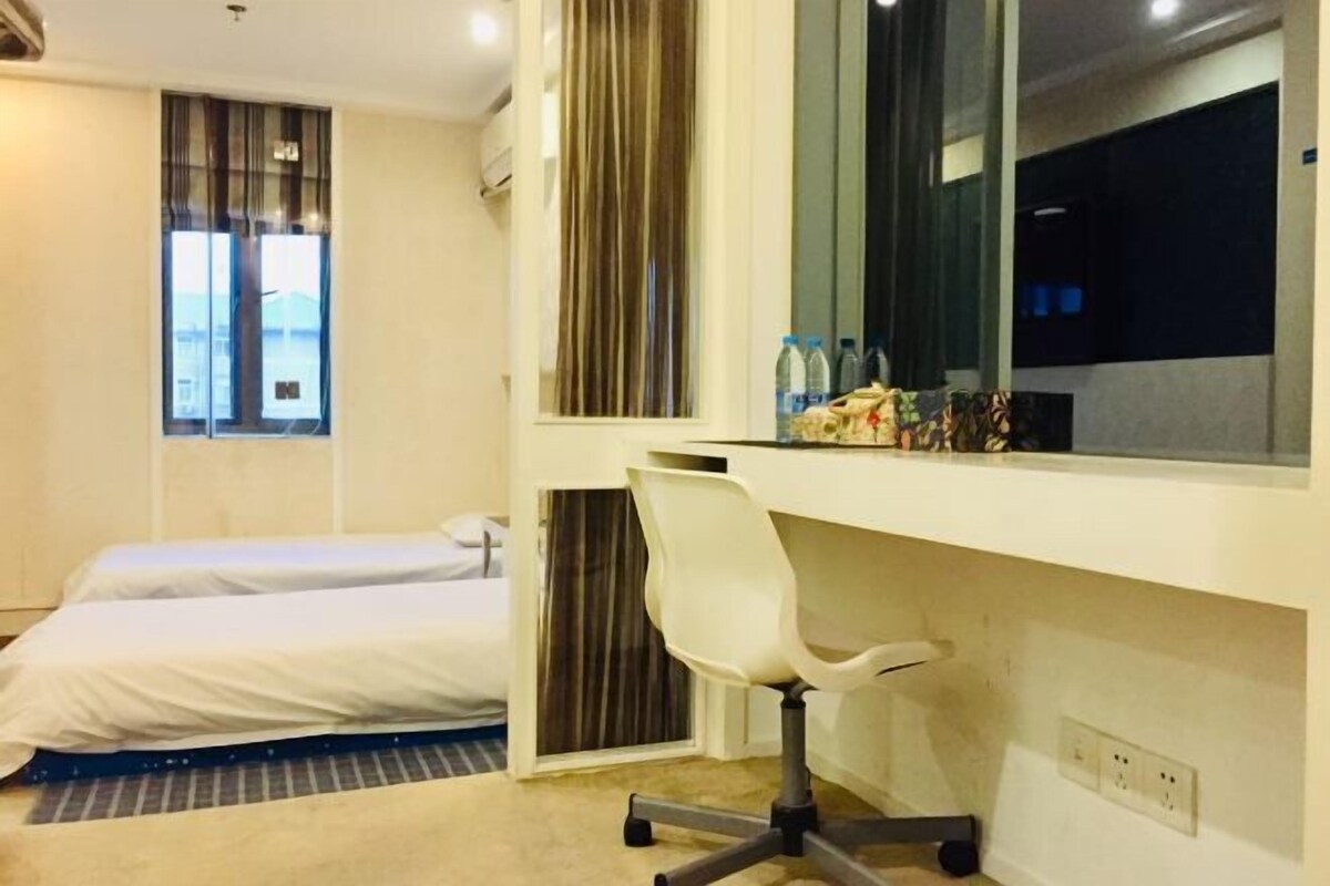 Quarto de hotel com cama, escrivaninha com cadeira, janela com cortinas e uma pequena mesa com garrafas.