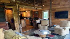 A cozy log cabin interior with a living room featuring a sofa and armchairs, a kitchen area, and a large TV mounted on the wall.